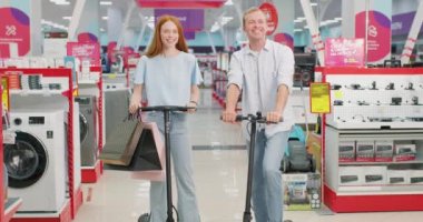 Home Electronics Store 'da mutlu çift scooter sürüyor. Erkek ve Kadın Güvenilir Çamaşır Aletleri Arayışında. Müşteriler Bir Perakende Dükkanında Modern Çamaşır Çözümlerini Keşfediyor