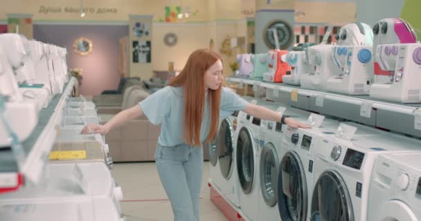 沉思沉思的困惑女人 长长的红头发疡洗衣机 打开洗衣机的门 打开洗衣机里的电器 女士在两台洗衣机之间进行选择 — 图库视频影像