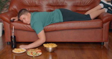 Obez çocuk kanepede uyuyor, uyanıyor, Coca Cola içiyor, tekrar uykuya dalıyor, şişko çocuk elinde bir tabak cips, burger, abur cubur, ağır çekim tembelliği olan çocuk pasif yaşam tarzını yönetiyor.