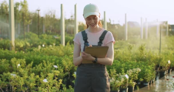 幸せな生姜の女性の庭師は植物の質をチェックし ノートブックにメモを取り 前衛的または温室に立って — ストック動画