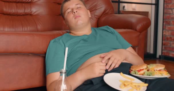 睡眠不足的胖少年吃饱了快餐后 躺在地板上 张开肚子 摸着自己的胃慢动作 — 图库视频影像