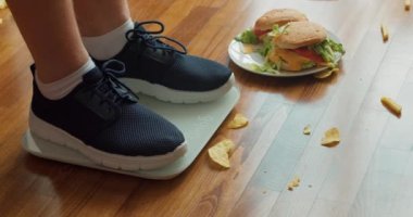 Spor ayakkabılı obez ergen evde kilo ölçeğinde duruyor, hamburgerler yerde ağır çekimde başarısız oluyor, fast food yemeyi reddediyor, vücut bakımını durduruyor, abur cuburu durduruyor, sağlıklı beslenmeyi düşünüyor.