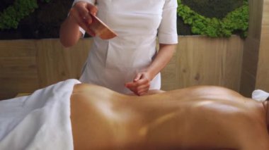 Kozmetik merkezindeki sarışın kadının sırtına aroma yağı döken masör. Aromaterapi masajı, ağır çekim sağlık ve vücut bakımı, masaj hazırlığı.