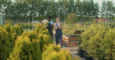 Kendine güvenen kızıl saçlı tarımcı kız ve Afrikalı Amerikalı iş arkadaşı serada yürüyor, konuşuyor, ağaçları işaret eden kadın çiçekçiye çiçek yetiştiriyor bitkilerle nasıl ilgilenileceğini anlatıyor.