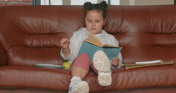 漂亮漂亮的小女孩拿着一本书 一边吃着苹果 一边坐在沙发上 慢动作 做家庭作业的女学生 — 图库视频影像