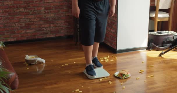 太った男の子は体重を量ろうとし 悪い結果について悲しいです ティーン手を振って 体重を失うことを拒否し 健康的な食べ物を食べる 弱い意志力 床の上で健康的でない食事や生活のジャンクフード — ストック動画