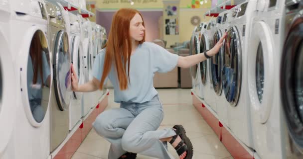 困惑伤心的顾客女孩无法决定买什么洗衣机 动作缓慢 垂头丧气的女孩手托着下巴坐在新洗衣机之间 — 图库视频影像