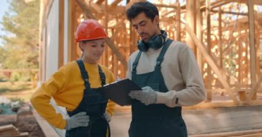 İnşaat alanında elinde dosya olan iki mühendis ağır çekimde çalışan erkek ve kadın mimarlar ellerinde kağıtlarla koruyucu miğfer takarak çalışma sürecini tartışıyorlar.