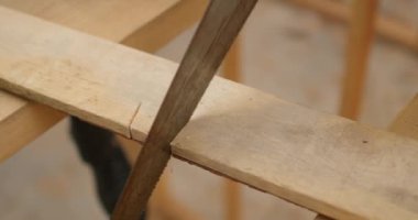Eldivenli marangoz bir işçiyle tahtayı testereyle kesiyor. Ağır çekimde metal testere kullanıyor. Sırada ev yapımı için gerekli aletler var.