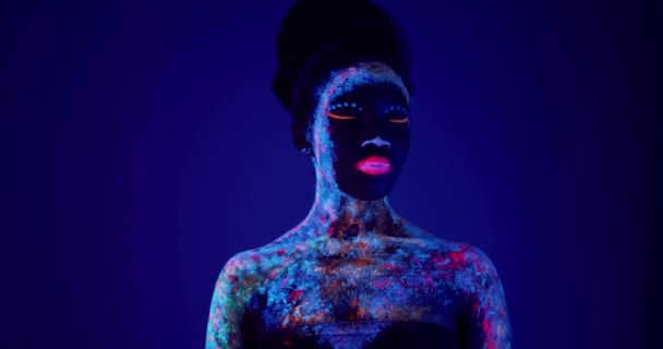 美丽的非洲裔美国女孩 脸上有紫外线图案 慢慢地摇了摇头 看着相机 优雅的造型 身体上有紫外线图案 在紫外线下发光的身体艺术 — 图库视频影像