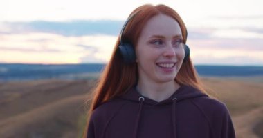 Uzun kızıl saçlı gülümseyen genç bir kadın dağın tepesinde durmuş mavi kapüşonlu müzik dinliyor, dinleniyor, dinleniyor, muhteşem manzaranın tadını çıkarıyor. Arka planda yavaş çekim mavi gökyüzü.