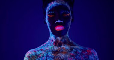Afro-Amerikalı, sakin sakin bir kız. Neon Makyajlı kameraya bakıyor, gözleri kapatıyor. Ağır çekim.