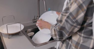 Ellerinde bulaşık takımı tutan, lavaboda su altında tabak yıkayan, köpüklü sarı sünger kullanan ve beyaz parlak bir mutfakta duran, ender görülen sırt manzaralı kız.