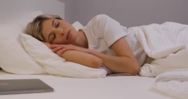 Genç sarışın kadın kolunu okşuyor, rahat bir yatakta uyuyor, gevşemiş sarışın kız yatak odasında dinleniyor. Yavaş çekim yorgunluğu gevşeme zamanı. Uyuma zamanı, dizüstü bilgisayar ön planda.