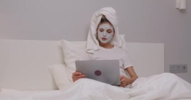 Yüzünde kozmetik maskeyle güzel bir genç kadın rahat bir yatakta uzanıp dizüstü bilgisayarla sohbet ederken ağır çekim kız internette geziniyor, boş zamanlarında internette geziniyor.