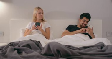 Genç evli çift akıllı telefon kullanıyor, kadın kıskançlıkla erkeğin telefonuna bakıyor. Güven yok. Çift ilişkileri, bağımlılık, neşeli adam telefonda sevgilisinden mesaj okuyor. Kadın erkeği rahat bırakıyor.