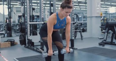 Spor kıyafetleri giyen, öne eğilip halterleri kaldıran, arka planda ekipmanları olan ciddi derecede motive olmuş bir kadın. Spor salonunda çalışan bir bayan sporcu. Geri çalışma Yavaş çekim kenar görünümü