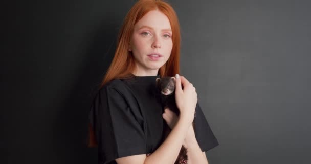 美丽的红头发女人抱着可爱的小雪貂 在工作室里摸着它 抱着它看相机 慢动作 爱情的概念 最好的朋友孤独的黑色背景 — 图库视频影像