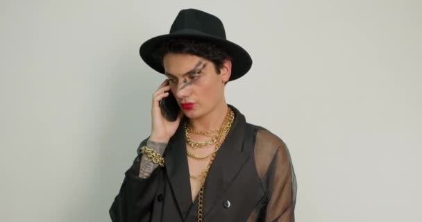 トランスジェンダーの男は 携帯電話のグレーバックグラウンドコミュニケーション黒ファッショナブルな衣装で豪華なアメリカのゲイ チェーンと帽子のアクセサリーで会話 Lgbt ライフスタイル — ストック動画