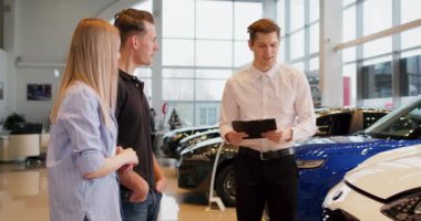 Yakışıklı satıcı müşterilere yeni bir araç gösteriyor. Yavaş çekim. Güzel mutlu çift araba galerisinde araba satın alıyor. Satıcıyla yeni otomobil satın alma konusunda aile danışmanlığı.