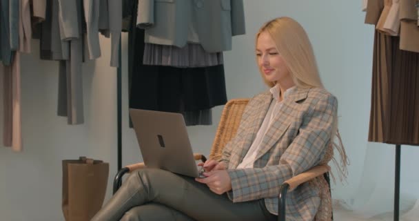 年轻的女时尚设计师在衣架附近的工作场所使用笔记本电脑 发短信 与顾客交流反对服装挂在衣架上慢动作在线个人造型 — 图库视频影像