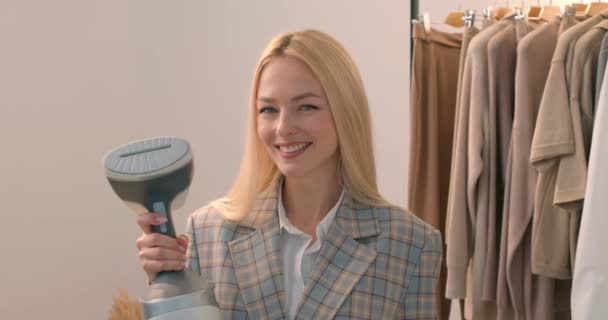 笑容可亲的漂亮金发女人手握手提式蒸汽机看着相机慢动作技术 家居服手持式熨烫机的必备器具 — 图库视频影像