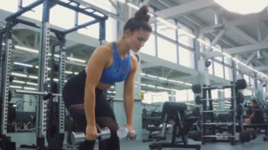 hırslı atletik çekici beyaz kadın spor giyimli iki kollu dambıl sırtı egzersizi, spor salonunda sırt, kol ve omuz kaslarını güçlendirme alıştırması yapıyor. Sağlık motivasyonu.