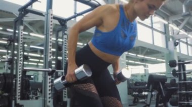 Motive olmuş, pantolonlu ve mavi bluzlu atletik kız spor salonunda halterlerle bükme egzersizi yapıyor. Kendine özen gösterme fitness vücut geliştirme konsepti. Sağlıklı yaşam tarzı, kamera yavaş çekim
