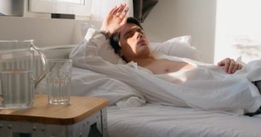 Mutsuz, susamış alkolik adam sabah yatağında su içiyor. Adam akşamdan kalma. Ağır çekim. Akşamdan kalma ya da migren hidrasyon konsepti.