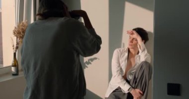 Genç adam sandalyede oturuyor yüzünü gün ışığından saklıyor, dizlerine sarılıyor, kadın fotoğrafçı bacaklarını yavaş çekimde fotoğraf çekiyor.,