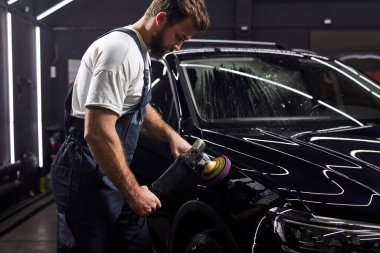 Yörünge parlatıcısı olan üniformalı bir adam tamir atölyesinde araba parlatıyor. Araba temizleme konsepti. İş yerinde sakallı yakışıklı bir adamın portresi, araba yüzeyine cilalayıcı sürüyor. Boşluğu kopyala