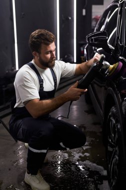 Kendine güvenen araba servisi çalışanı yörünge cilası kullanıyor, oto tamirhanesinde siyah araba cilalıyor, üniformalı adam otomobille ilgileniyor, temizlik yapıyor.