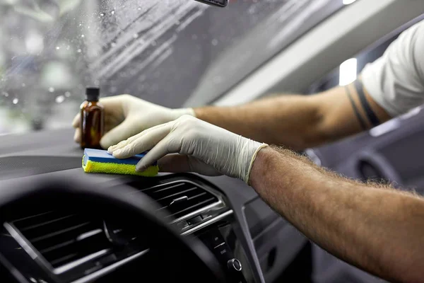 Auto repair man in protective gloves cleaning auto from inside using detergent oils liquid from bottle applied on rag sponge, side view, close-up hands of cropped male