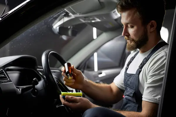 professional car wash service worker cleaning car interior with special chemical anti dust liquid. side view on male holding bottle with liquid in hands applying it on rag