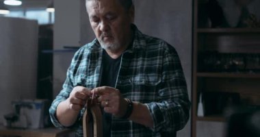 Başarılı, olgun bir zanaatkar deri ve deri malzemelerden yapılmış eşyaları restore eder. Sakallı Asyalı adam atölyede dikiş dikmeye odaklanmış. Ciddi profesyonel bir zanaatkar iğneyi delikten geçirir.