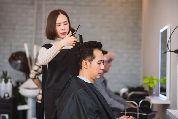 Asian woman hairstylist using scissors and comb for hair cut and design on man\'s hair in barbershop.