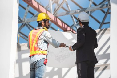 İnşaat alanında takım çalışanları, güvenlik kaskı takan iki inşaat mühendisi dijital tablet ve taslak kullanarak fabrikada çalışırken....