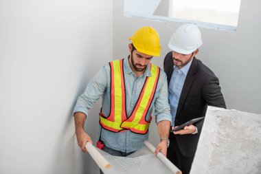 İnşaat alanında takım çalışanları, güvenlik kaskı takan iki inşaat mühendisi dijital tablet ve taslak kullanarak fabrikada çalışırken....