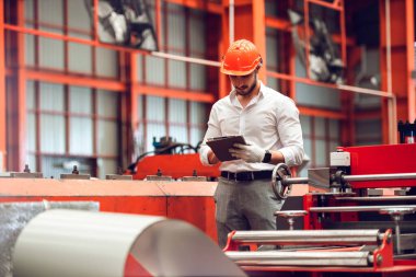 Fabrikadaki makine sistemini denetlemek için tablet kullanan koruyucu kask takan fabrika işçisi mühendisi. Heavy metal endüstrisi üretim fabrikası.