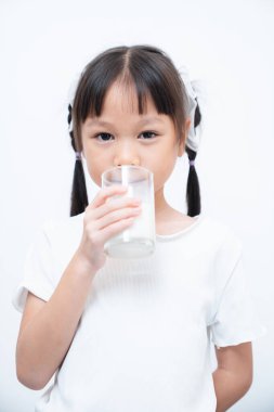 Genç Asyalı bir kızın portresi taze süt içerken, elinde beyaz bir arka planda bir bardak tutarken, çocukluk beslenme ve süt ürünlerinin faydalarını en az düzeyde ve temiz bir şekilde vurguluyor.