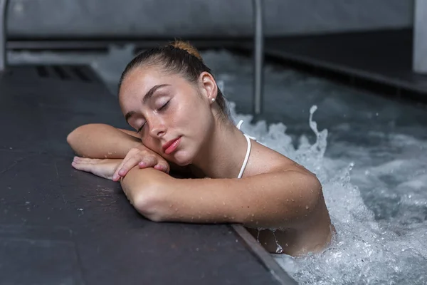Calm female folding arms on pool border and closing eyes while resting in bubbling water of pool in spa