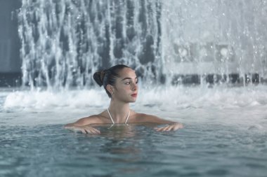 Young brunette looking away and swimming in clean pool near fountain while spending weekend day on spa resort