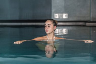 Sakin, genç bir bayan, saçlarını topuz yapıp gözlerini kapatıyor ve dinlenme yerindeki spa seansında havuzda yüzüyor.