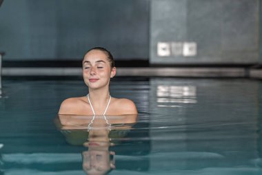 Pozitif genç bir bayan, hafta sonu kaplıcada temiz havuz suyunda dinlenirken gözlerini kapayıp gülümsüyor.