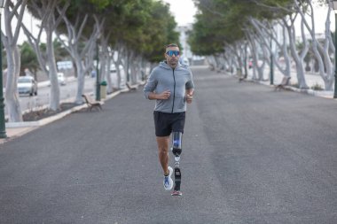 Tam vücut güçlü, protez bacaklı olgun erkek sporcu yaz günü parkta kardiyo eğitimi sırasında asfalt yolda koşuyor.