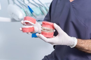 Tanımlanamayan tıbbi üniformalı erkek dişçi ve lateks eldiven. Modern ışık kliniğinde otururken takma dişleri ve diş tellerini gösteriyorlar.