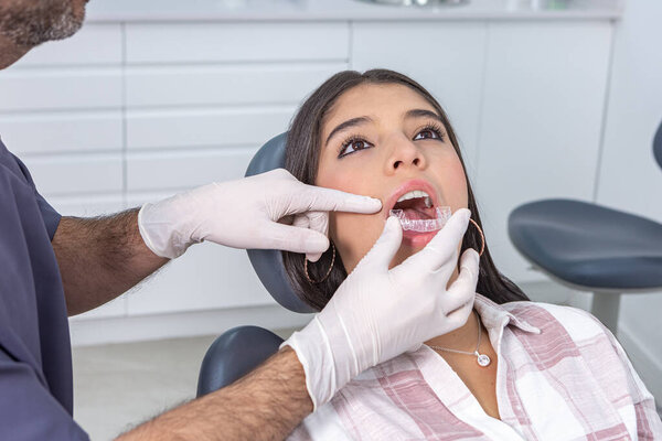 Crop unrecognizable adult bearded male dentist in uniform and gloves trying transparent vacuum formed retainer on teeth of ethnic female teenager during treatment in clinic