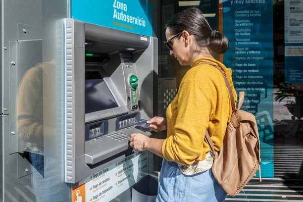 Woman wearing sunglasses and carrying a backpack on her back withdrawing money with a credit card at a automatic cash dispenser