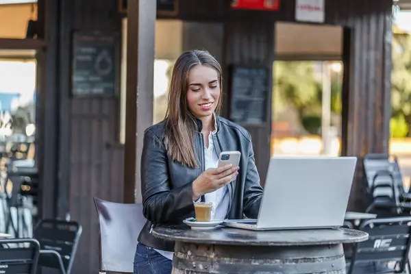 Positive young female entrepreneur in casual outfit sitting at round table with netbook and cup of hot coffee while chatting on cellphone during remote work
