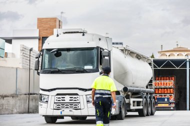 Güneşli bir günde fabrikada iş saatinde tanker kamyonunun yanında yürüyen üniformalı ve şapkalı isimsiz erkek makine mühendisi.